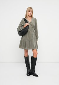 Olivengrønn, langermet kjole laget av myk stoff med knyteliv og flagrende hem. Båret med høye svarte støvler og en svart skulderveske.