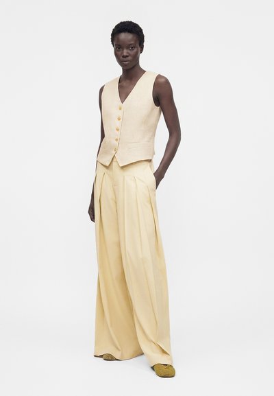 Γυναίκα που φορά αμάνικο μπεζ γιλέκο με κουμπιά και φαρδιά πλισέ κρεμ παντελόνια με κίτρινα πλεκτά παπούτσια, στέκεται με το ένα χέρι στην τσέπη.