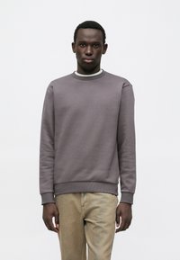 Sweatshirt gris à encolure ronde, manches longues et poignets côtelés, porté avec un pantalon beige. Le tissu semble doux et texturé.