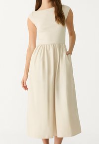 Beige dress with a fitted, ribbed top and a flowing skirt; features cap sleeves, an elastic waistband, and side pockets.