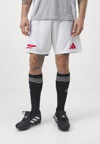 Pantalones cortos de fútbol blancos con el logotipo rojo de Arsenal, combinados con calcetas negras con franjas plateadas y zapatos de fútbol negros con acentos blancos.