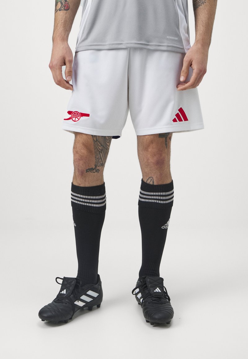 Pantalones cortos de fútbol blancos con el logotipo rojo de Arsenal, combinados con calcetas negras con franjas plateadas y zapatos de fútbol negros con acentos blancos.