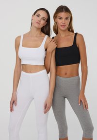 Due modelle stanno fianco a fianco, una indossa un crop top bianco a scollo quadrato e leggings bianchi abbinati, l'altra un crop top nero e leggings grigi.