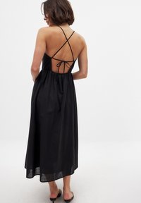 Musta, selkä avoin mekko, jossa on ohuet ristikkäiset hihnat ja solmittava yksityiskohta takana. Laskostettu, joustava vyötärö ja polven puoliväliin asti ulottuva pituus. Tasainen tekstuuri.