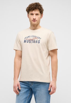 Junger Mann mit lockigem Haar, der ein beiges T-Shirt mit der Aufschrift "Jeans Company Mustang" und blaue Jeans trägt, steht vor einem einfarbigen Hintergrund.