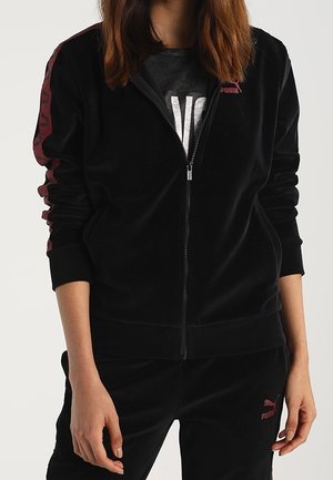 Kobieta ubrana w czarny dres Puma z bordowymi logo i paskami, kurtka na zamek częściowo rozpięta, ukazująca ciemną koszulkę pod spodem.