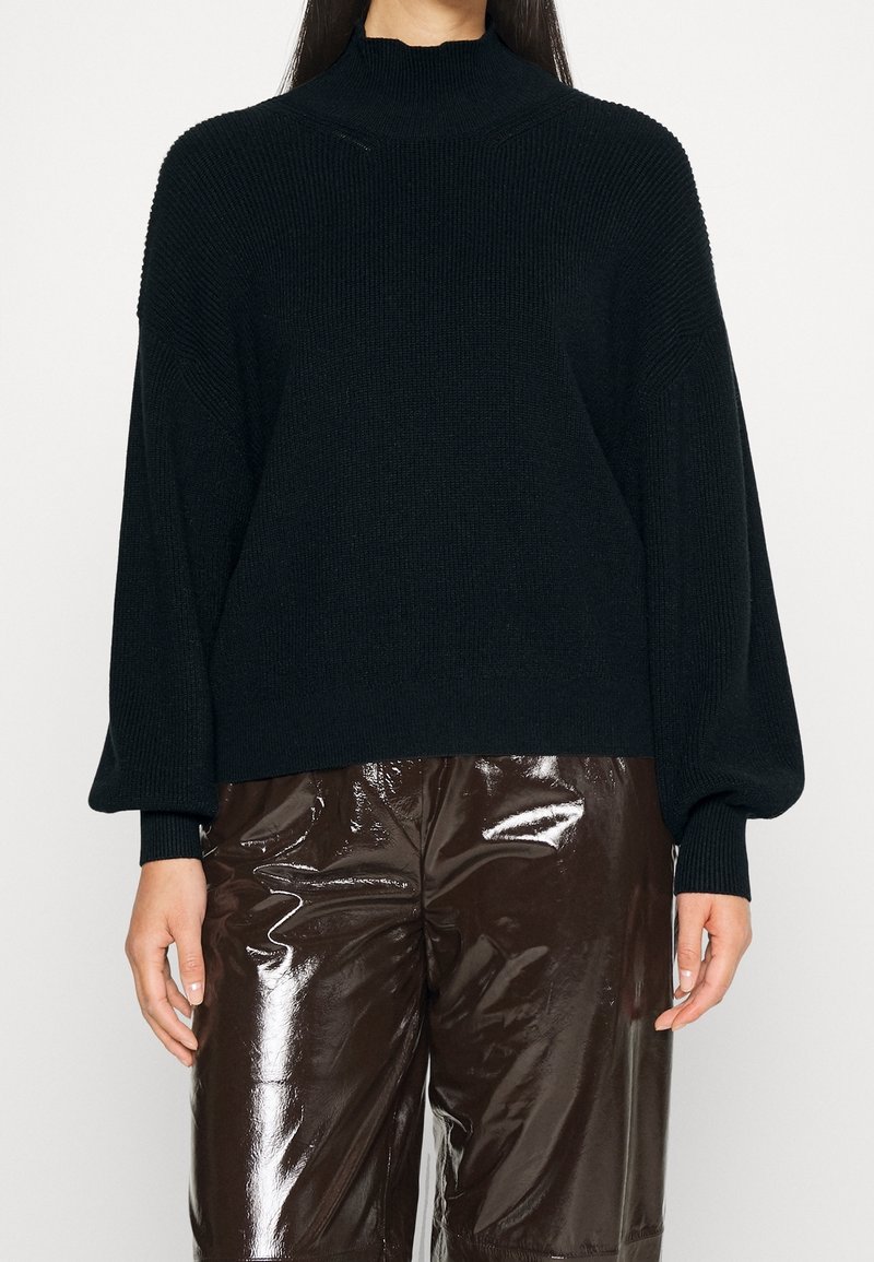 Maglione nero lavorato a maglia con colletto alto, spalle cadenti e texture a coste. Abbinato a pantaloni in pelle sintetica lucida marrone.