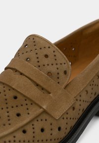 Καφέ δερμάτινα μοκασίνια από σουέντ με διάτρητες λεπτομέρειες και φαρδιά λουράκι στη φάπα. Μαύρη εξωτερική σόλα, λεία εσωτερική επένδυση για άνεση.