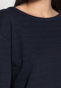 Vrouw in een donkerblauwe, gestructureerde gebreide trui met een brede ronde hals en lange mouwen.