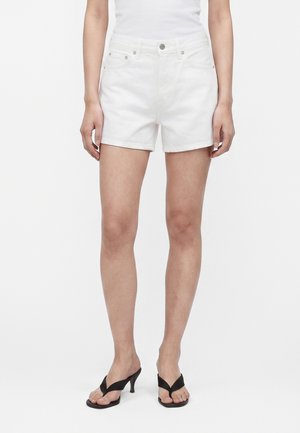 Kvinde iført hvide denimshorts, hvid top og sorte sandaler med hæl står mod en ensfarvet hvid baggrund.