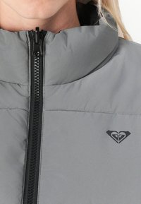 Primo piano di una giacca a vento grigia con zip, colletto alto e logo del marchio a forma di cuore nero sul petto.