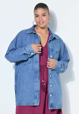 Persona con cabello corto que lleva pendientes de aro, anillos plateados, un vestido morado y una chaqueta vaquera azul de gran tamaño sobre un fondo blanco.