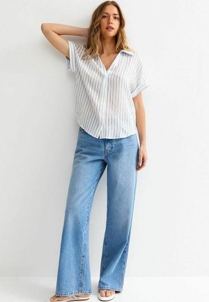 Femme debout, un bras levé, portant une chemise blanche à manches courtes à boutons avec des rayures bleues et un jean bleu clair à jambes larges.