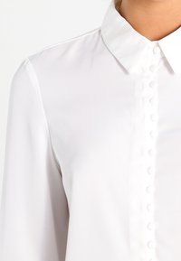 Camicia bianca con colletto a punta e bottoni rivestiti in tessuto, indossata da una persona visibile dal collo fino alla parte centrale del torso.