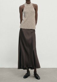 Beige gestricktes Tanktop mit hohem Hals, kombiniert mit einem fließenden braunen Satinrock. Schwarze Schuhe mit Riemchen-Detail runden den Look ab.