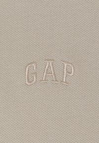 Svijetlo bež pamučna tkanina s teksturiranim pletenim uzorkom. Sadrži bež izvezeni natpis "CAP" centriran na tkanini.