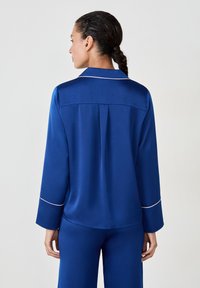 Haut de pyjama en satin bleu avec col, manches longues et passepoil blanc le long des poignets et du col. Dispose d'un pli dans le dos pour faciliter le mouvement.