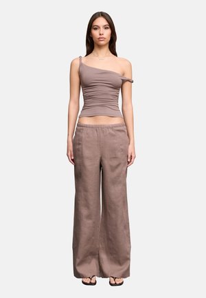 Junge Frau, die frontal steht und ein mauvefarbenes, einseitiges, gerafftes Top, weite Hosen und schwarze Sandalen vor weißem Hintergrund trägt.