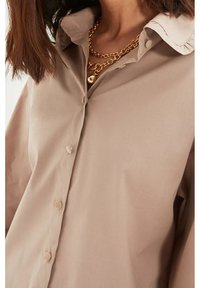 Chemise beige à boutons avec un col classique, présentant une texture lisse et des boutons visibles le long de la patte de boutonnage, collier en chaîne dorée superposé.
