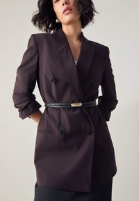 Dwurzędowa marynarka z ciemnofioletowego materiału z czarnymi guzikami, z podwiniętymi rękawami i czarnym paskiem ze złotym akcentem.