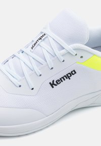 Baskets blanches de sport avec une tige en mesh, featuring un logo "Kempa" noir, des lacets bleu clair et un accent jaune près du talon.