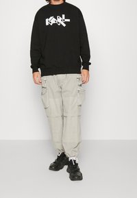 Czarna bluza z kapturem z białym logo „KARL”, zestawiona z jasnoszarymi spodniami typu cargo z licznymi kieszeniami i masywnymi czarnymi sneakersami.