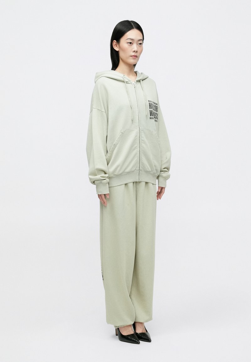 Lichtgroene zip-up hoodie met grote voorzakken, gecombineerd met bijpassende wijde broek. Opvallend gedrukt logo op de borst; gladde textuur.