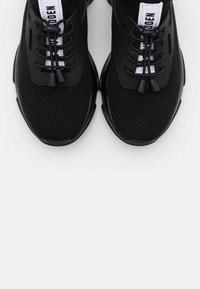 Paire de baskets de sport noires en tissu mesh, lacets élastiques noirs sans nouer, et étiquettes de languette blanches avec texte noir sur fond blanc.