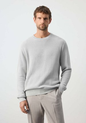 Hombre con cabello corto y castaño vistiendo un suéter de punto gris claro y pantalones beige claro, de pie con una mano en el bolsillo sobre fondo blanco.