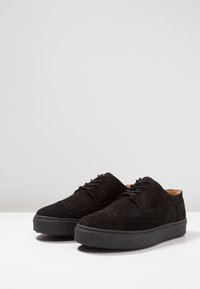 Černé semišové boty s plochou gumovou podrážkou, kulatou špičkou a dekorativním broguem. Obsahují šněrování a minimalistický design.