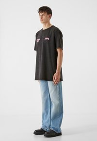 Črna oversized majica s rožnatim napisom na prsih, kombinirana s svetlo modrimi širokimi hlačami. Manekenka nosi črne debelodne čevlje.