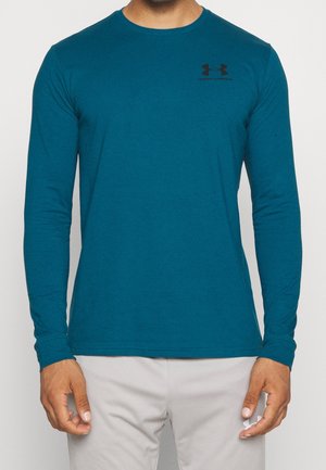 Mann mit einem langärmeligen tealfarbenen Under Armour-Shirt und hellgrauer Hose, steht vor einem einfarbigen Hintergrund.