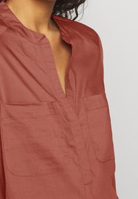 Chemise légère couleur rouille avec un col pointu, un décolleté en V et deux poches poitrine. Texture froissée qui ajoute de la profondeur au design.