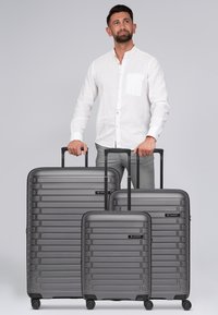 Trois valises rigides gris foncé de tailles différentes avec des rainures horizontales et des poignées télescopiques, dotées de roues et d'accents noirs.