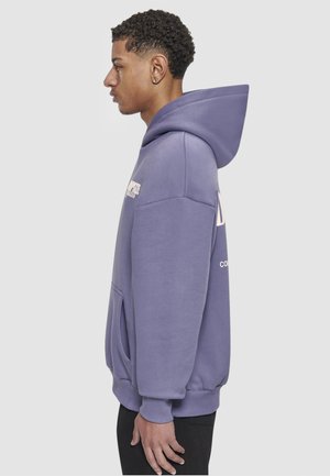 Noor mees lilla kapuutsiga dressipluusis seisab profiilis, käed lõdvestunult külgedel, taust on ühevärviline.