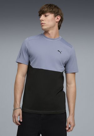 Młody mężczyzna ubrany w krótkim rękawem, dwukolorową niebiesko-czarną koszulkę Puma, patrzący w lewo na jednolitym szarym tle.