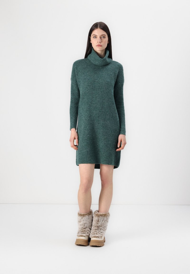 Green knitted dress with a high, folded collar and long sleeves, featuring a relaxed fit and soft texture. Paired with furry, brown ankle boots.