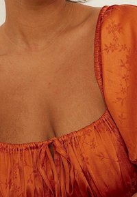 Blusa de seda naranja con un patrón floral texturizado, escote elástico y detalle de lazo en la parte delantera, con mangas abullonadas.