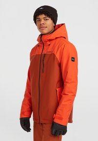 Orange og brun skijakke med lynlås foran, sidelommer og justerbar hætte. Sorte handsker og hue komplimenterer outfittet.