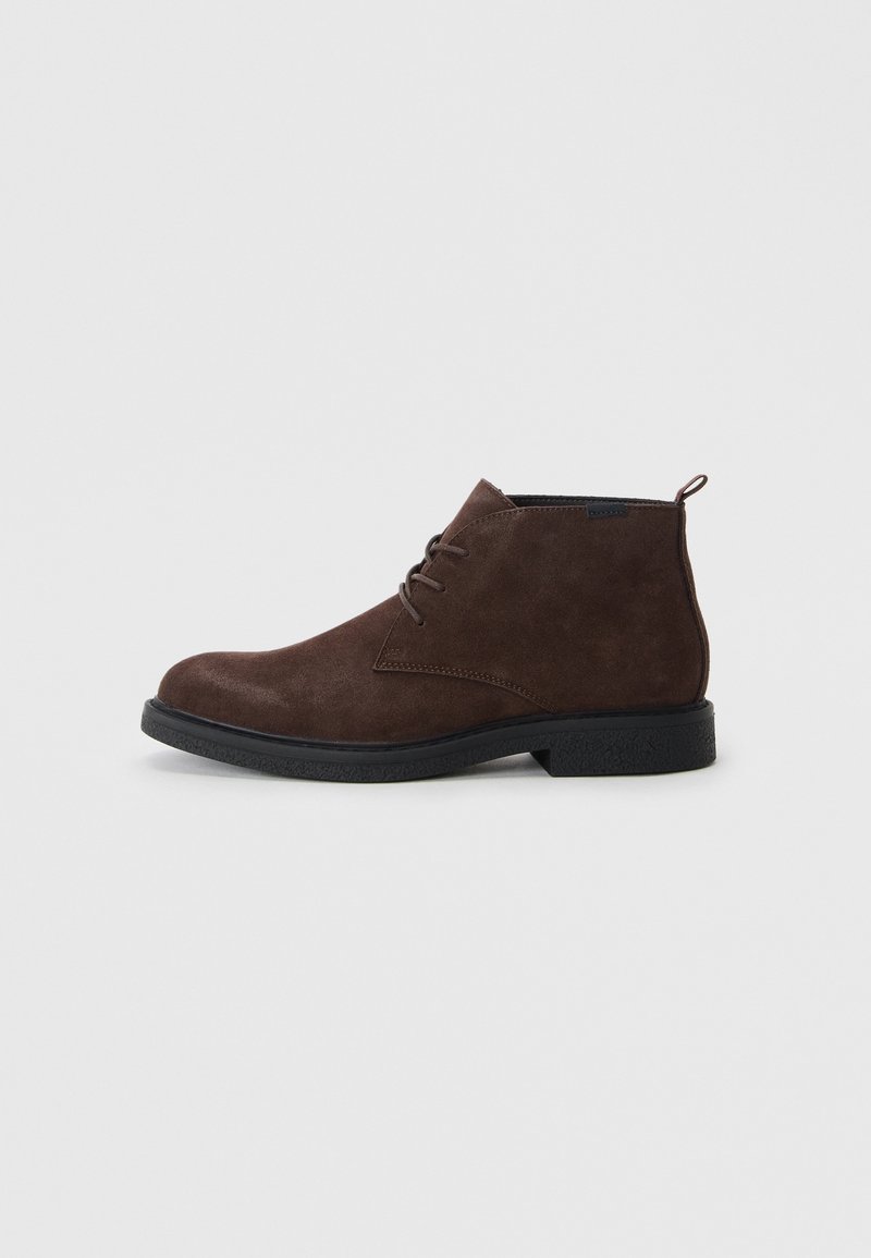 Bottine en daim marron avec semelle en caoutchouc noire et fermeture à lacets à trois œillets, présentée de profil sur un fond gris clair.
