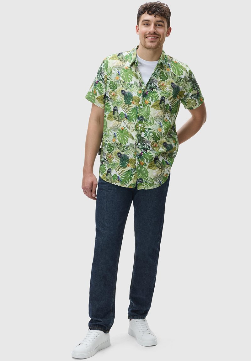 Camicetta verde a maniche corte con motivo di foglie tropicali, abbinata a jeans blu scuro e sneaker bianchi, in posa in piedi su uno sfondo chiaro.