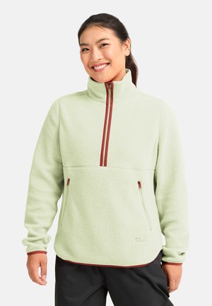 Lachende vrouw met een lichtgroene fleece trui met bordeaux ritsdetails en zwarte broek, staand tegen een effen achtergrond.
