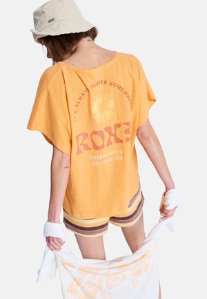 Pomarańczowa koszula z krótkim rękawem z kwiatowym nadrukiem i napisem na plecach, zestawiona z pasiastymi szortami w odcieniach ziemi. W dłoni biała ręcznik.