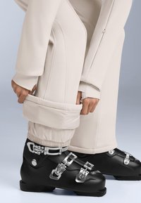 Czarne buty narciarskie z srebrnymi elementami, noszone z beżowymi, ocieplanymi spodniami narciarskimi. Spodnie mają elastyczne mankiety i gładką teksturę.