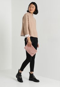 Suéter de punto beige con mangas anchas, jeans skinnies negros, botas negras de tobillo con detalles de hebilla y un bolso de mano de cuero rosa con solapa.