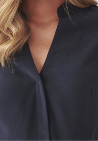 Marineblauwe blouse met een V-hals en zonder kraag. Gemaakt van een gestructureerde stof met een gladde uitstraling en een subtiele glans.