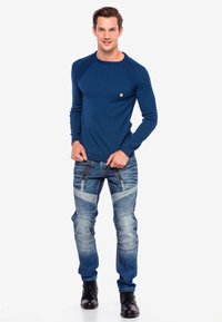 Marineblaues langärmliges Shirt mit Rippstruktur, kombiniert mit blauen, verwitterten Jeans mit diagonalen Details, Reißverschlussakzenten und schwarzen Schnürstiefeln.