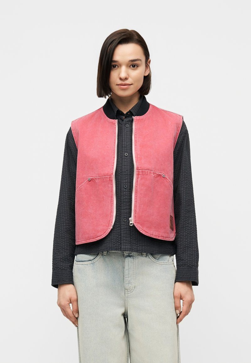 Νεαρό άτομο με ίσια μαύρα μαλλιά φορά ροζ γιλέκο με φερμουάρ πάνω από μαύρο πουκάμισο με κουμπιά και ανοιχτό μπλε τζιν, στέκεται μπροστά σε λευκό φόντο.