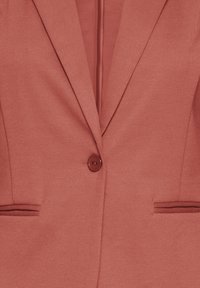 Blazer rose avec une texture lisse, fermeture à un bouton, et deux poches, présentant un col classique.