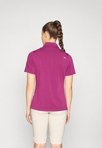 Lila Poloshirt mit kurzen Ärmeln, glatte Textur, mit kleinem Logo auf der Rückseite. Kombiniert mit hellen Shorts.